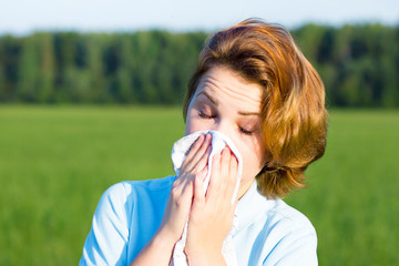 Young woman blowing her nose with tissue, snot in paper handkerchief outdoor. Sick girl sneezes  in a summer field, park. Natural background. Feeling unwell, ill, bad, suffering. Allergy