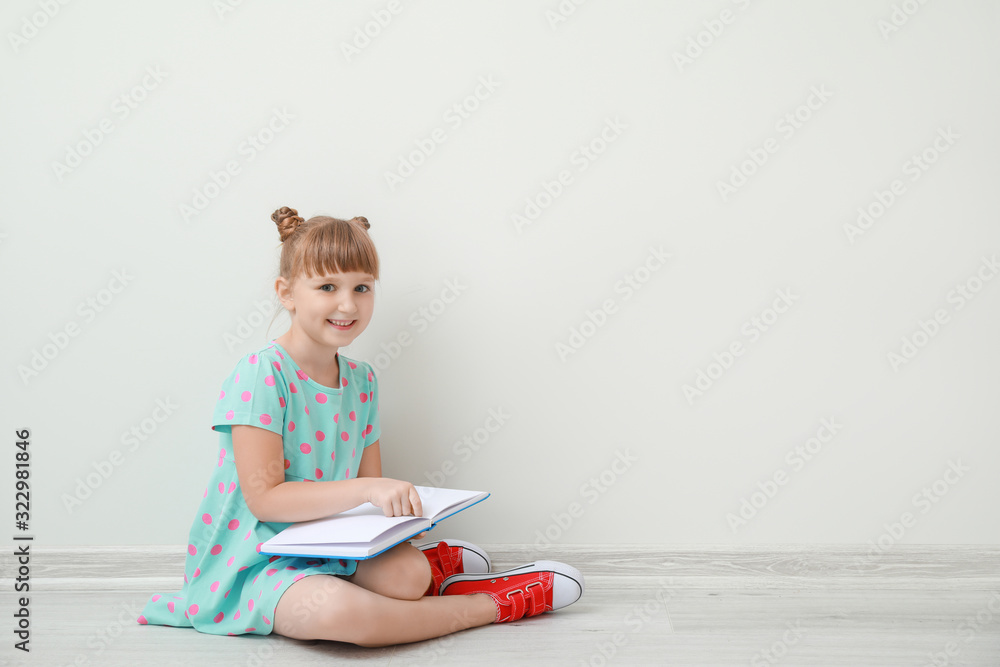 Wall mural Cute little girl with book sitting near white wall