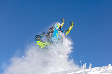 elite sports snowmobiler rides and jumps on steep mountain slope with swirls of snow storm. background of blue sky leaving a trail of splashes of white snow. bright snowmobile and suit without brands