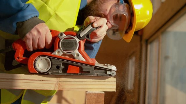 SLOW MOTION, CLOSE UP, DOF: Young Caucasian builder with a beard is sanding a wooden beam at a busy construction site. Experienced contractor uses an electric sander to smooth out the edge of a plank.