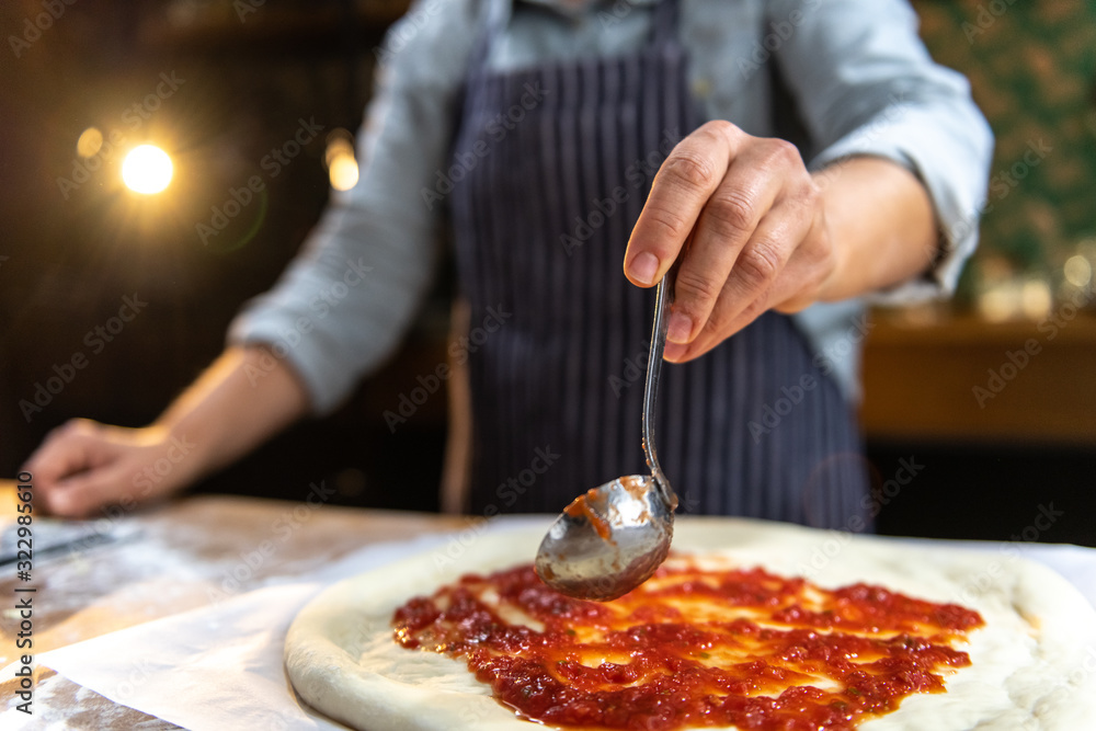 Wall mural female chef is pouring freshly made tomato sauce over a traditional, homemade pizza.