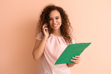 Young African-American businesswoman with documents talking by phone on color background