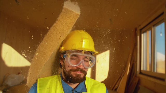SLOW MOTION, CLOSE UP, PORTRAIT, DOF: Funny shot of a smiling builder getting hit with a thick piece of foam. Bearded male surveyor wearing a yellow helmet gets struck in the head by a piece of foam.