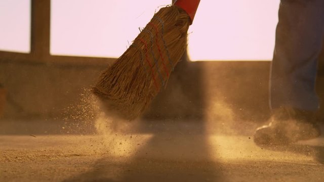 SLOW MOTION, CLOSE UP, LOW ANGLE: Unrecognizable worker sweeps the dusty floor after a long day at a busy construction site. Contractor is sweeping the dirty floor with a straw broom at sunset.