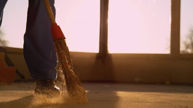 SLOW MOTION, CLOSE UP, LENS FLARE, LOW ANGLE: Contractor is sweeping the dirty floor with a retro straw broom at sunset. Worker sweeps the dusty floor after a long day at a busy construction site.