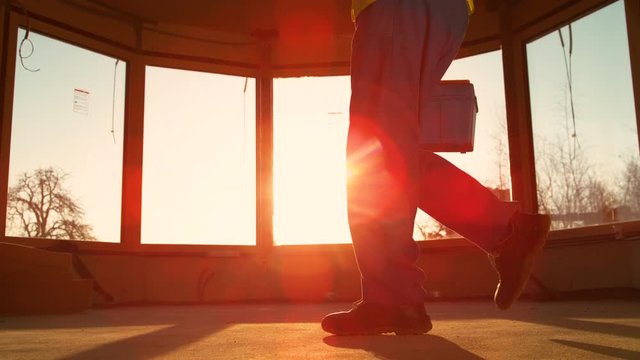 SLOW MOTION, LOW ANGLE, CLOSE UP, LENS FLARE: Builder coming to work carries his toolbox across the room at sunrise. Unrecognizable man working at a construction site of a house leaves at sunset.