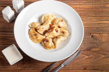 Dumplings and ravioli stuffed with meat. Sprinkle sliced smoked tomatoes. Served on a large white plate. Next to a bowl of sour cream. On a wooden background.
