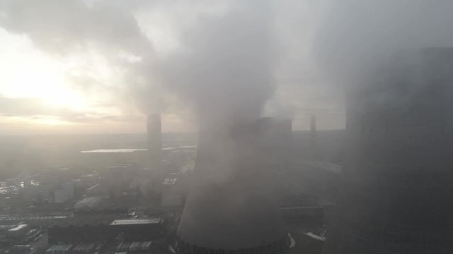Aerial push in view of UK power station cooling towers flying through smoke steam emissions at sunrise.
