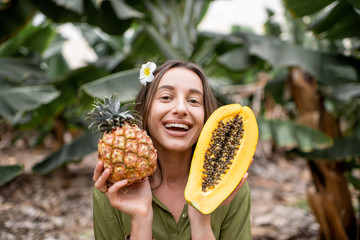 Portrait of a young smiling woman with sliced papaya and ananas outdoors on the tropical plantation. Concept of vegetarianism, healthy eating of fresh fruits and wellbeing - obrazy, fototapety, plakaty