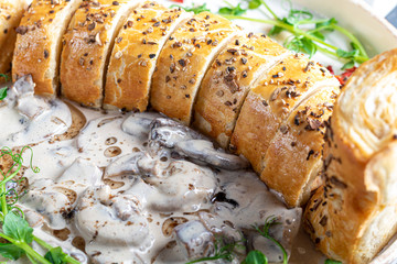 Puff pastry roll stuffed with mushrooms. Nearby are champignons in sour cream, sliced. Served with microgreen and sun-dried tomatoes. On a dark background.
