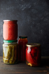 Preserved vegetables in jars, ont a dark background. Cucumbers, bell pepper, adjika, tomato juice