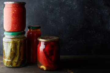 Preserved vegetables in jars, ont a dark background. Cucumbers, bell pepper, adjika, tomato juice