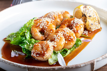 Shrimps cooked in teriyaki sauce with a slice of fried lemon on a pillow of lettuce leaves. On a white background, on a dark wooden background.