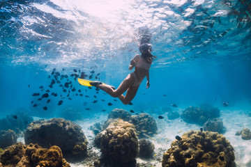 Woman glides underwater with yellow fins over sandy sea and coral.