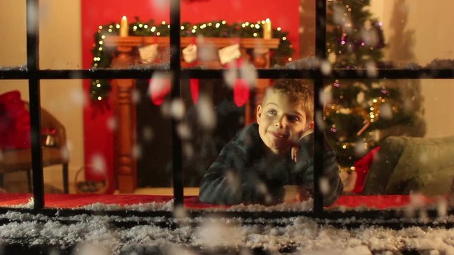 Child looking through Window at Christmas. Snow falling outside. Christmas Tree and fireplace in lounge