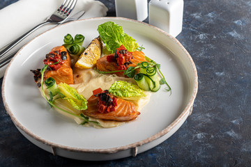 Celery and potato puree with pieces of red fish, garnished with herbs and fresh cucumbers. On a white plate with lemon slices. On a wooden table.