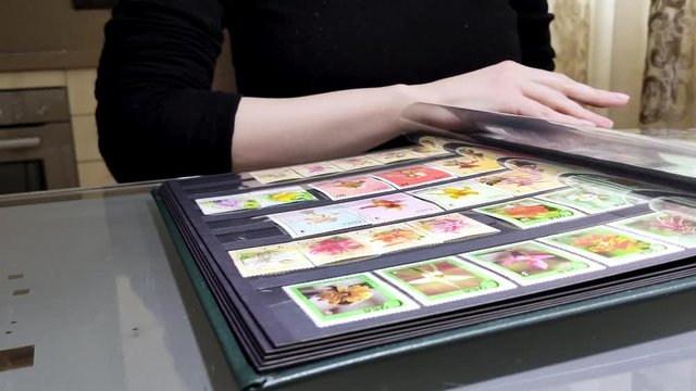 White girl sits at table in front of open album with collection of postage stamps. Woman in black sweater leafs through book with postmark. Philately hobby. Close-up side view