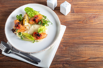 Celery and potato puree with pieces of red fish, garnished with herbs and fresh cucumbers. On a white plate with lemon slices. On a wooden table.