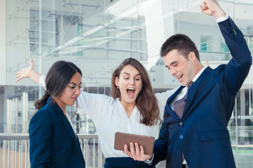 Three good looking colleagues standing in office corridor, man and woman in white blouse exulting, receiving good news, attractive woman in spectacles looking uncertain. Work, communication concept
