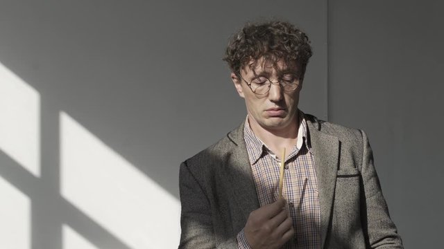 Man with glasses, with curly hair, in gray jacket, in white room with shadow from window on the wall, walks from side to side, holds pencil to his lips and reflects on another project or business idea