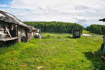 Obraz premium .Old ruined buildings in the village, rickety street wooden toilet on the background of beautiful nature, lake, forest, hills. Lush green grass, cloudy weather, white dog on a leash...