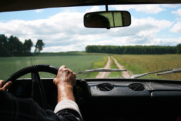 .Male tanned hands with a tucked shirt on the steering wheel of a riding car and the road, clouds, forest and field in front.