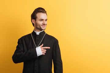 smiling catholic priest looking away and pointing with finger isolated on yellow
