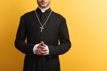 Cropped view of catholic priest standing with clenched hands isolated on yellow - obrazy, fototapety, plakaty