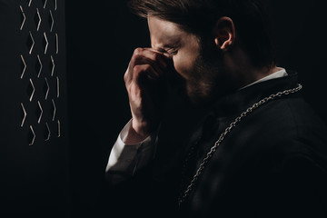 young tense catholic priest touching face with closed eyes near confessional grille in dark with...