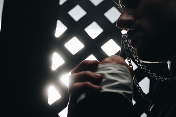 cropped view of catholic priest kissing cross on his necklace near confessional grille in dark with...