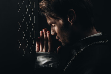 young catholic priest praying with closed eyes near confessional grille in dark with rays of light - obrazy, fototapety, plakaty