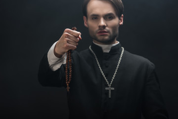 Selective focus of young confident catholic priest holding wooded rosary beads in outstretched hand...