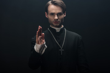 young confident catholic priest looking at camera and showing blessing gesture isolated on black