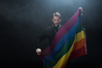 confident catholic priest holding lgbt flag while looking at camera on black background with smoke