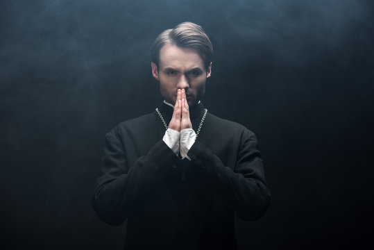 serious catholic priest looking at camera while praying on black background with smoke