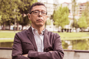 Serious middle-aged man posing at camera and standing in park. Guy standing with his arms crossed, green lawn, trees and buildings in background. Man portrait concept. Front view.