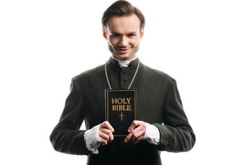 young, smiling catholic priest showing holy bible isolated on white
