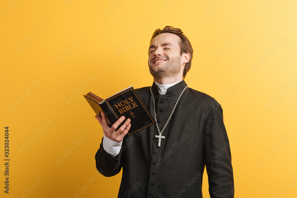 Wall mural excited catholic priest laughing while holding holy bible isolated on yellow - Wall murals