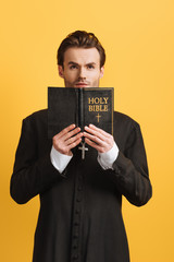 thoughtful catholic priest looking up while holding open bible isolated on yellow