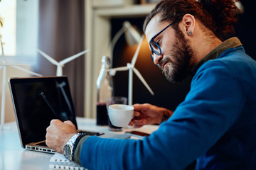 Young innovative dedicated bearded employee sitting in his modern office and drawing sketches of windmills and drinking coffee. Sustainable development concept.