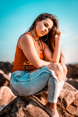 Lifestyle, look of a pretty brunette in a red shirt and jeans on a sunset sitting on a rock