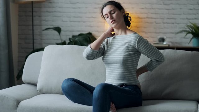 Video of young woman with back and neck pain sitting on the sofa in the living room at home.