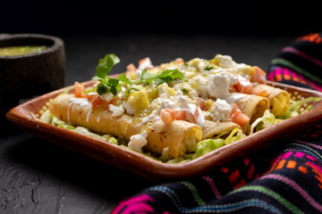 Mexican potato and cheese fried tacos also called flautas with green sauce on dark background