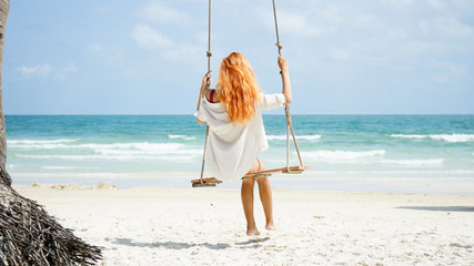 Beach summer vacation tropic palm style portrait of young beautiful girl on beach swing blue sea.Red haired woman swinging on the beach on Phu Quoc island, Vietnam. Happy  on tropical palm tree swing.