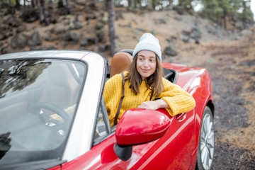 Portrait of a stylish woman in yellow sweater sitting on the driver's seat of convertible sports car on the roadside. Carefree lifestyle and traveling concept