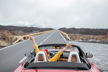 Young woman traveling by convertible car on the picturesquare road on the volcanic valley, raising...