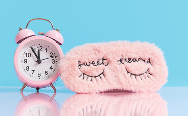 Sleep optimization. Pink fluffy mask for sleeping and a pink alarm clock on a light blue background.