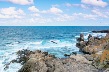 rocky sea shore in the afternoon. blue waves crashing the coast. fluffy clouds above the horizon.