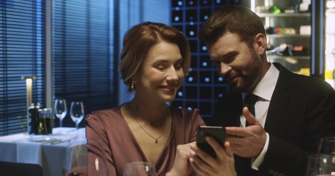 Caucasian good looking couple siting in restaurant in evening and watching something on smartphone. Married man and woman with phone in hand talking and discussing something at dinner.