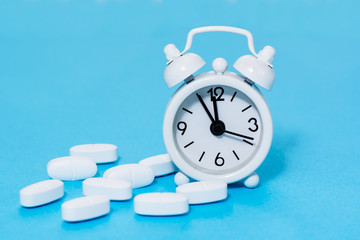 White alarm clock and white oval pills next to it on a blue background. Taking medicine at the right time.